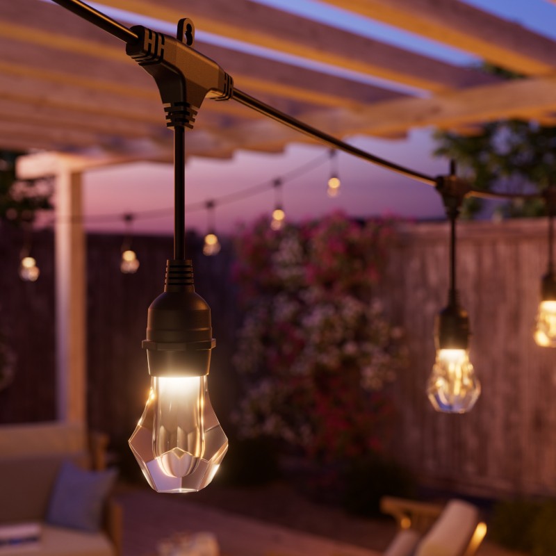 Close up of two warm white outdoor string lights LED bulbs hanging from a wooden patio. Wooden fence with flower vines in the background, and additional string lights hanging in the surroundings.