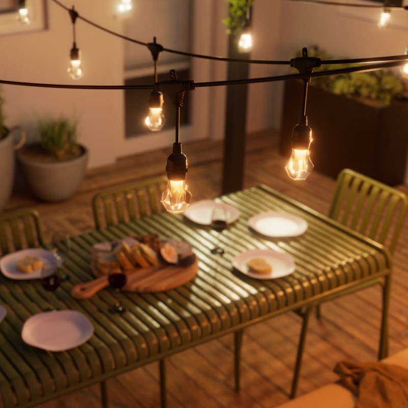 Cozy outdoor patio with BBQ grill, table, and outdoor string lights casting a warm yellow glow. A charcuterie spread on the table with plates set out, surrounded by plants